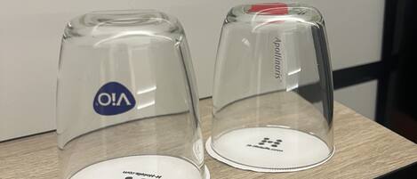 Two clear glass coffee cups with lids on a wooden table against a white wall.