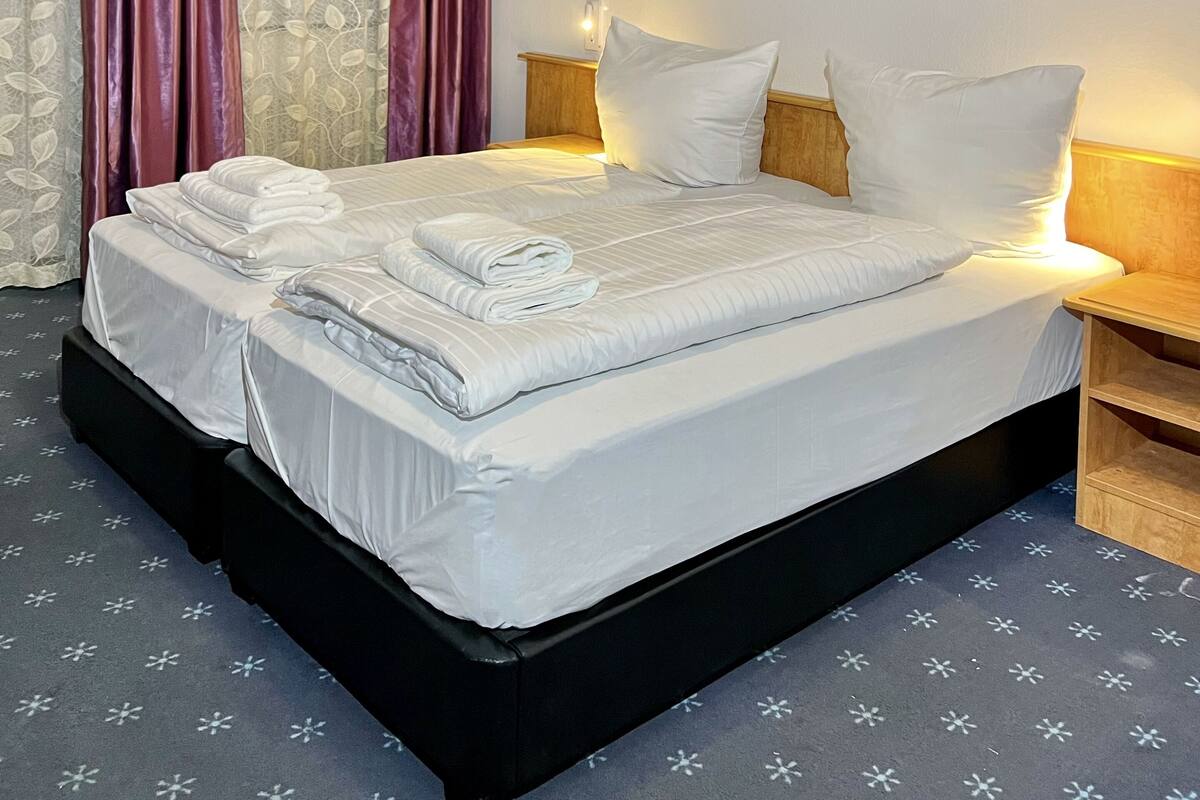 Een netjes opgemaakt bed met witte lakens en kussens, een nachtkastje met een lamp en een aan de muur bevestigd kunstwerk waarop een vaas met bloemen staat afgebeeld.