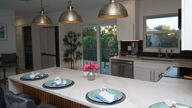 A modern kitchen with a marble countertop, pendant lights, and a dining area with a vase of flowers. - The Capri House: Fun, Grill & Beach Minutes Away (Palm Beach Gardens)