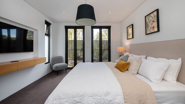 A modern bedroom with a large bed, a TV mounted on the wall, a chair, and a window with a view of trees. - Mister Montague by Property Mums (South Melbourne)