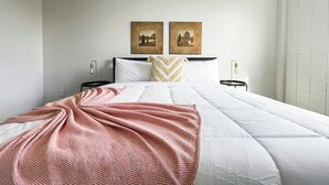 Una cama bien tendida con una colcha rosa y blanca, dos mesitas de noche con lámparas y dos cuadros en la pared.