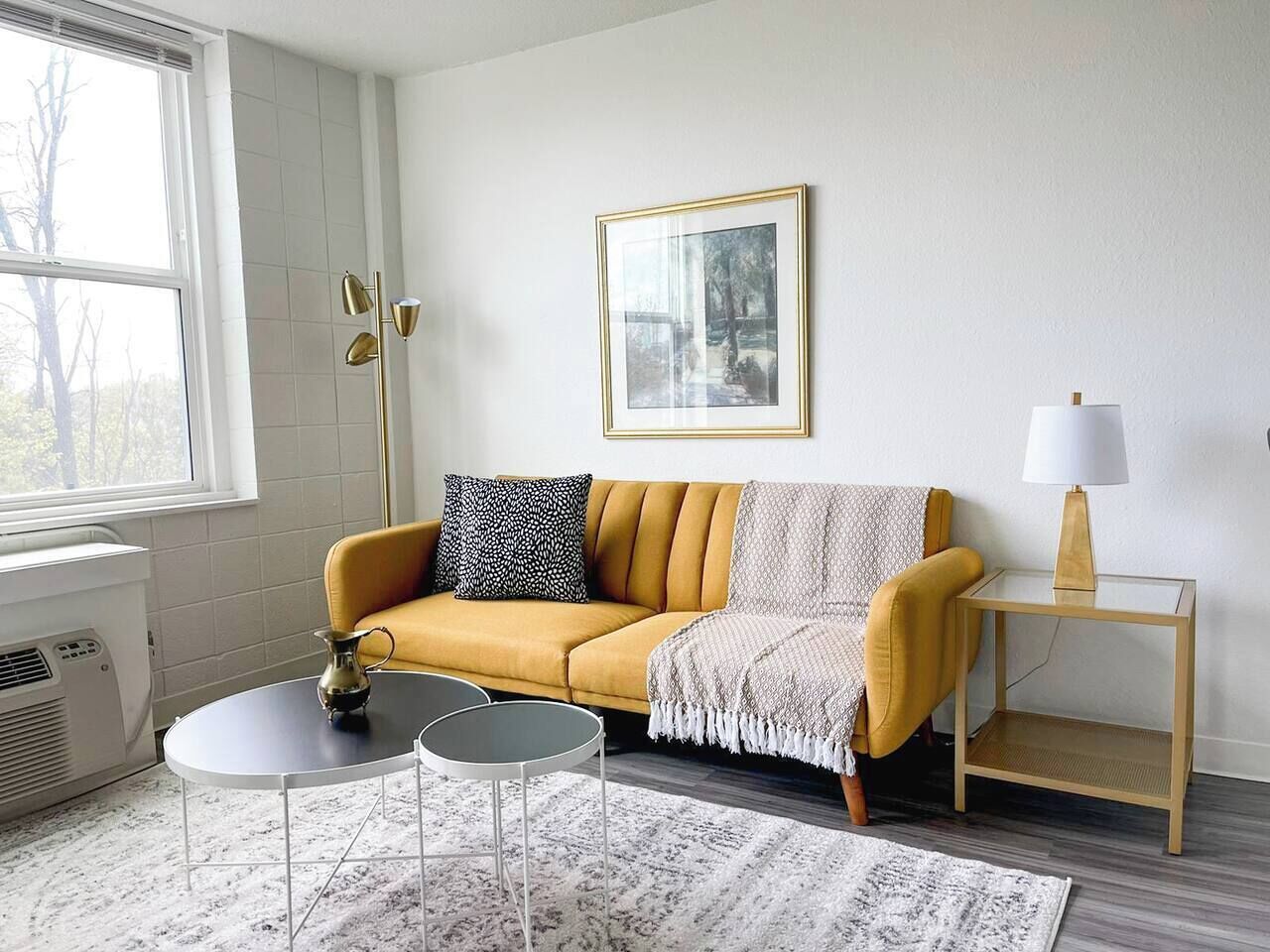 En stue med en sennepsgul sofa, et lille rundt bord, en lampe og et billede i ramme på væggen.