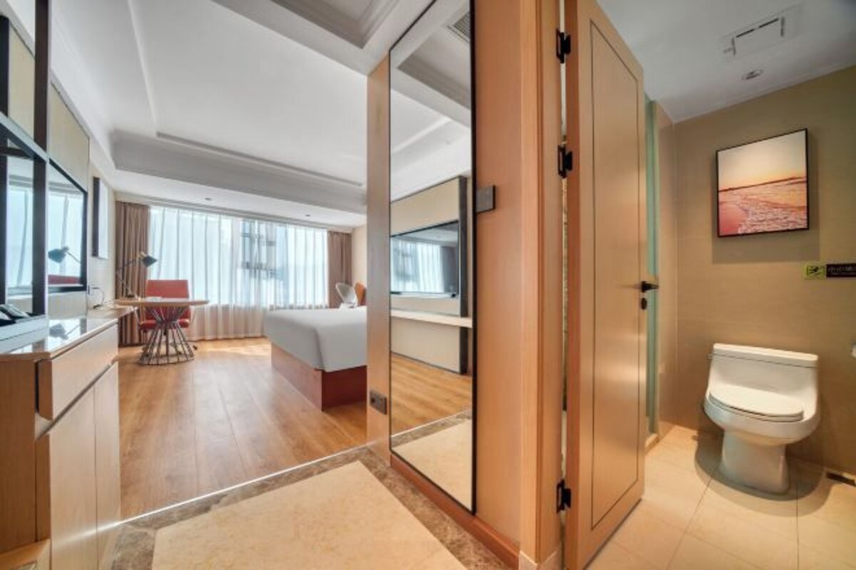 Ett hotellrum med en säng, ett skrivbord, en stol, ett badrum med toalett och ett stort fönster.