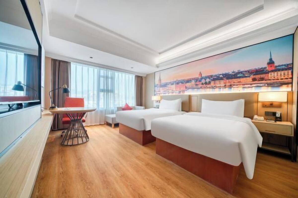 Habitación de hotel con dos camas, un escritorio, una silla, una mesa y un cuadro grande con vistas a la ciudad.