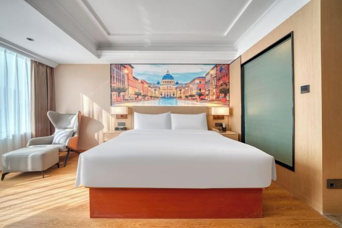 A hotel room with a large bed, a chair, a desk, and a painting of a cityscape on the wall.