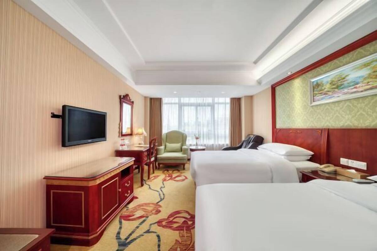 Habitación de hotel con televisión, escritorio, dos camas, una silla y ventana con cortinas.
