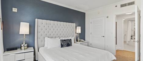 A neatly arranged bedroom with a tufted headboard, white bedding, two bedside tables with lamps, and a door leading to a bathroom.