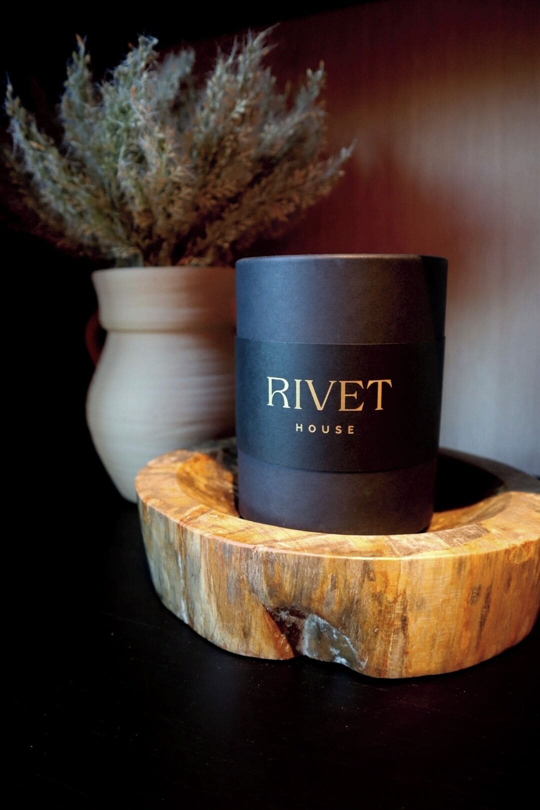 A black candle with "Rivet House" text on a wooden coaster.