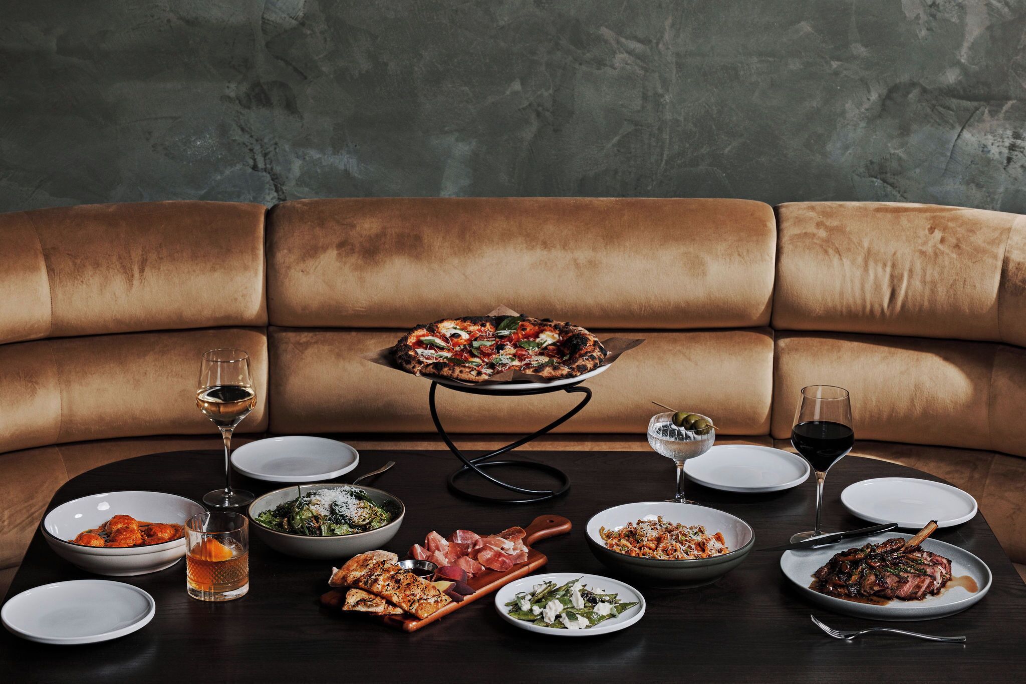 A dining table set with various dishes, glasses of wine, and a pizza on a stand.