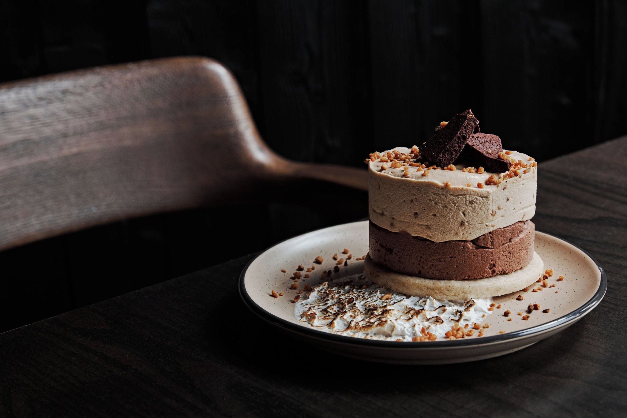 A layered dessert on a plate with crumbs and chocolate pieces on a dark wooden table.
