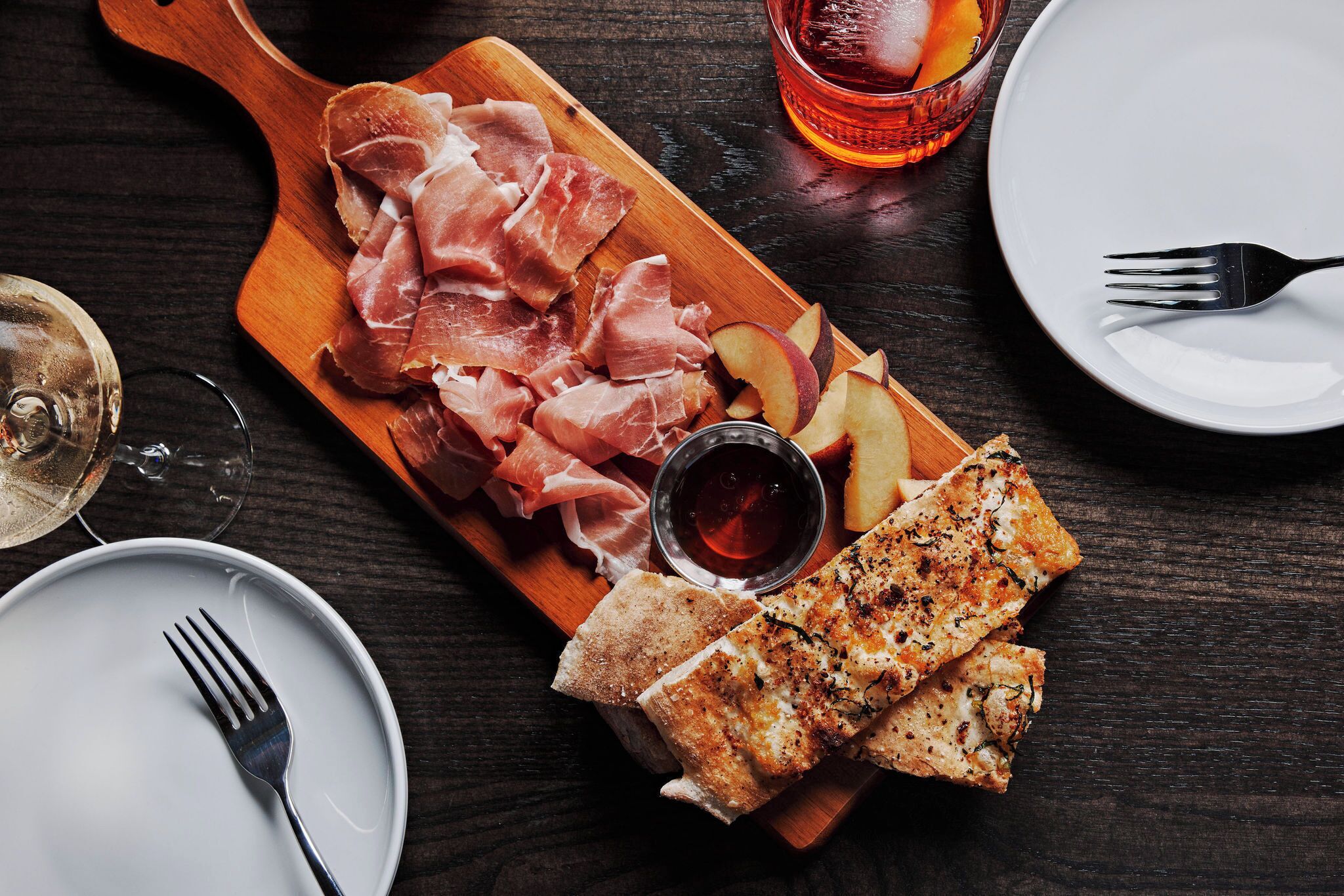 A wooden board with sliced meats and cheese, a glass of red drink, and a plate with a fork.