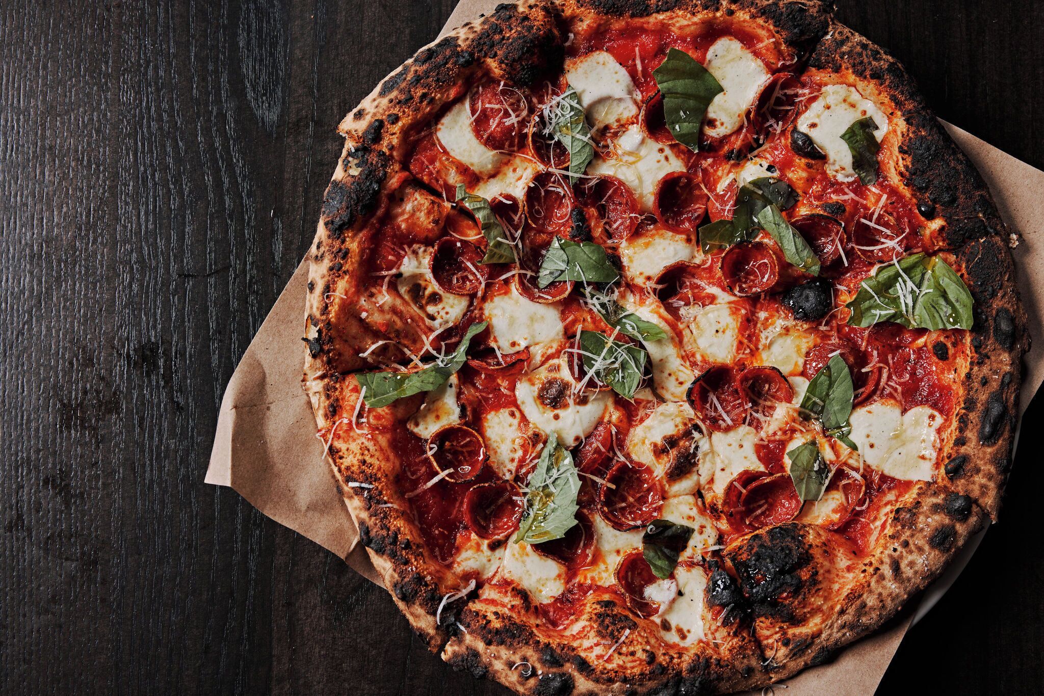Pizza con pepperoni, queso y albahaca sobre una superficie de madera oscura.