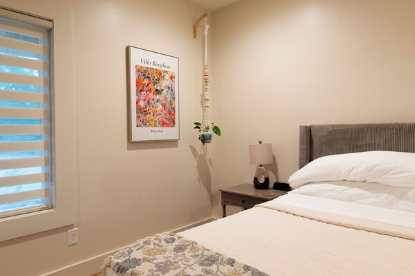 A bedroom with a bed, a nightstand, a lamp, a framed picture, and a window with blinds.