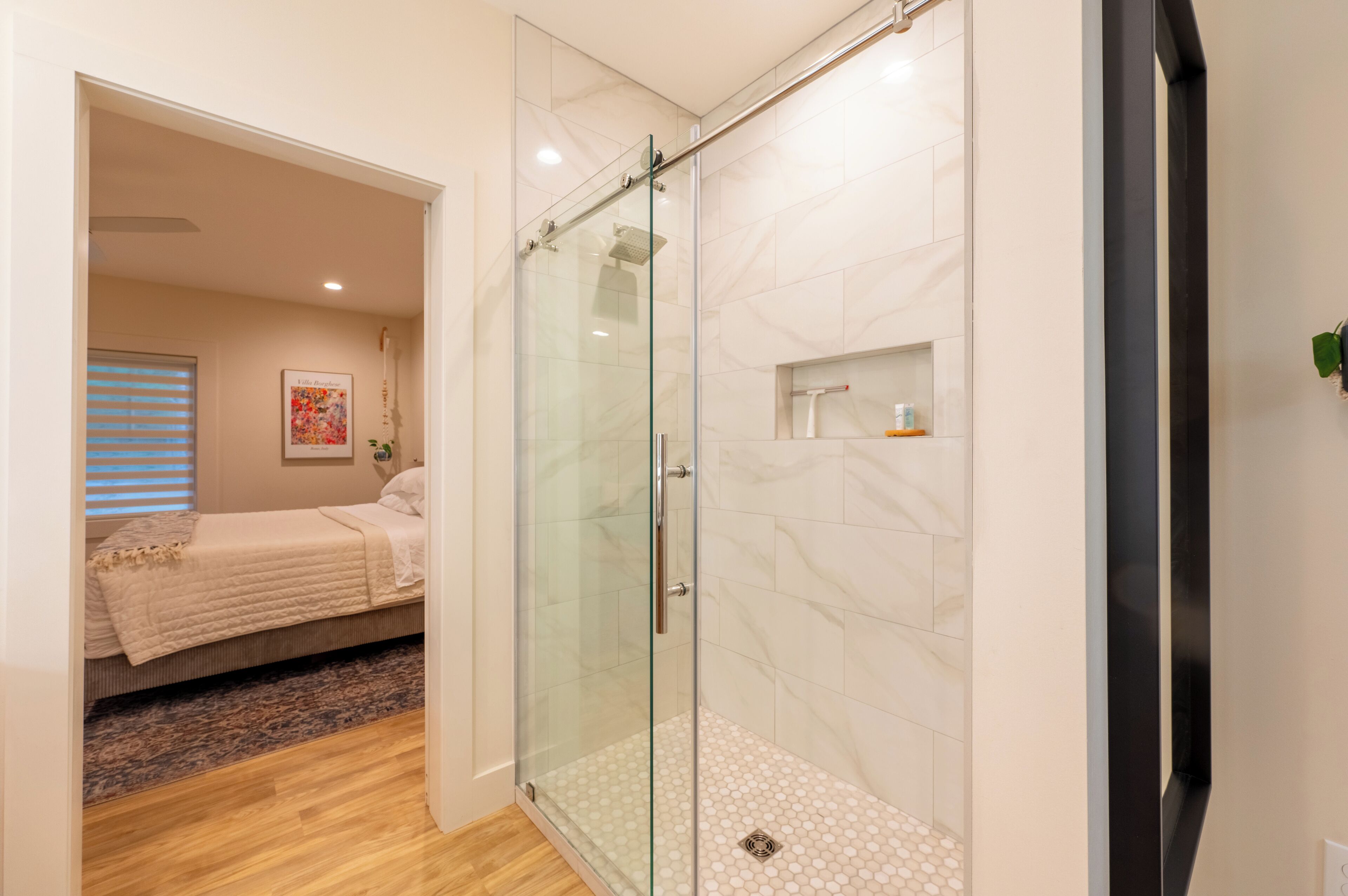 A modern bathroom with a glass shower enclosure, a wooden floor, and a bed in the adjacent room.