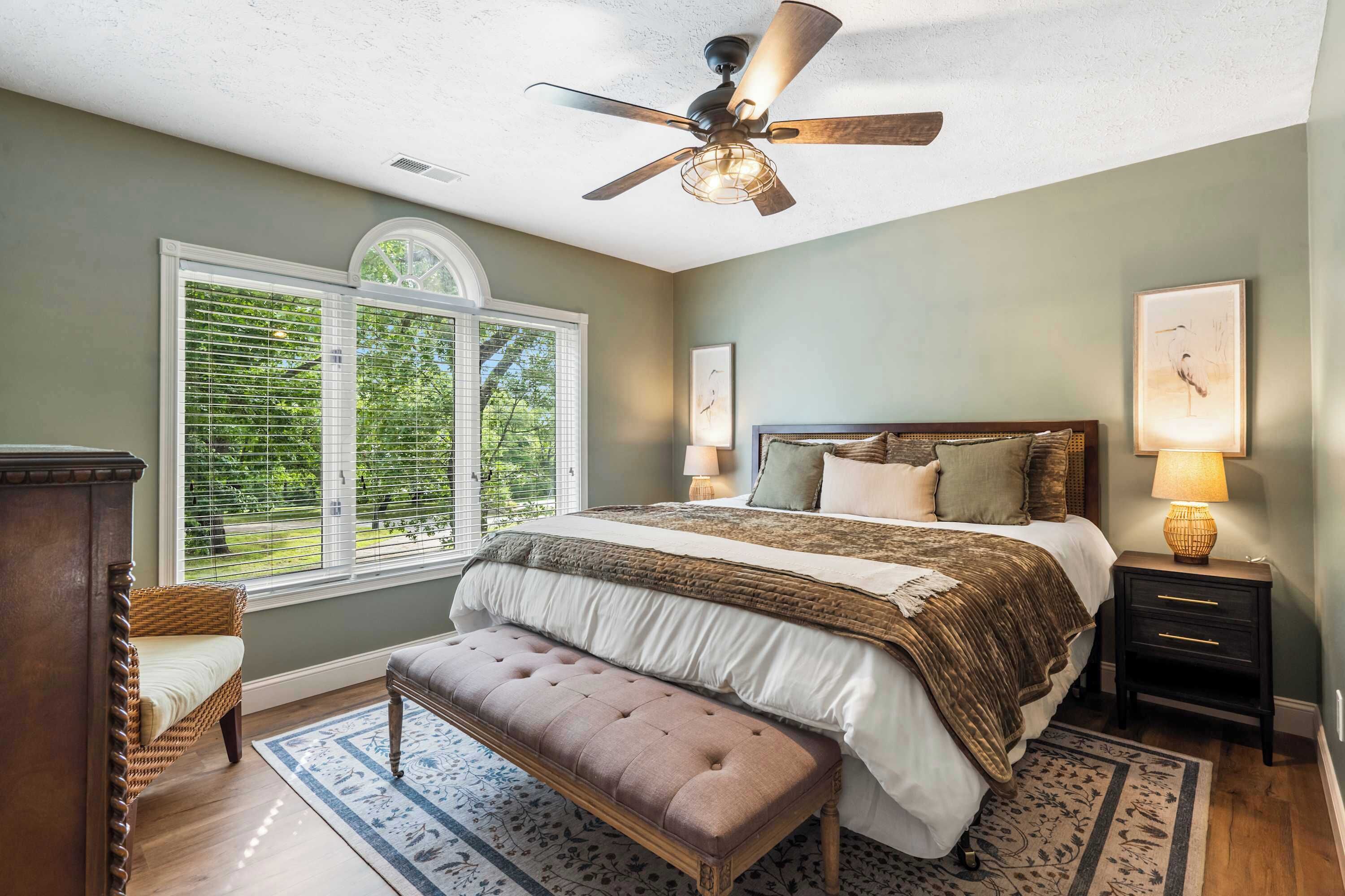 A bedroom with a large bed, a bench, a ceiling fan, a window with blinds, and a nightstand with a lamp.