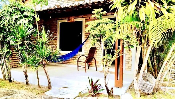 A rustic building with a brick exterior, a wooden door, and a hammock. There are several potted plants and a wicker basket near the entrance. - CHALÉS CENTRO DA PIPA (Tibau do Sul)