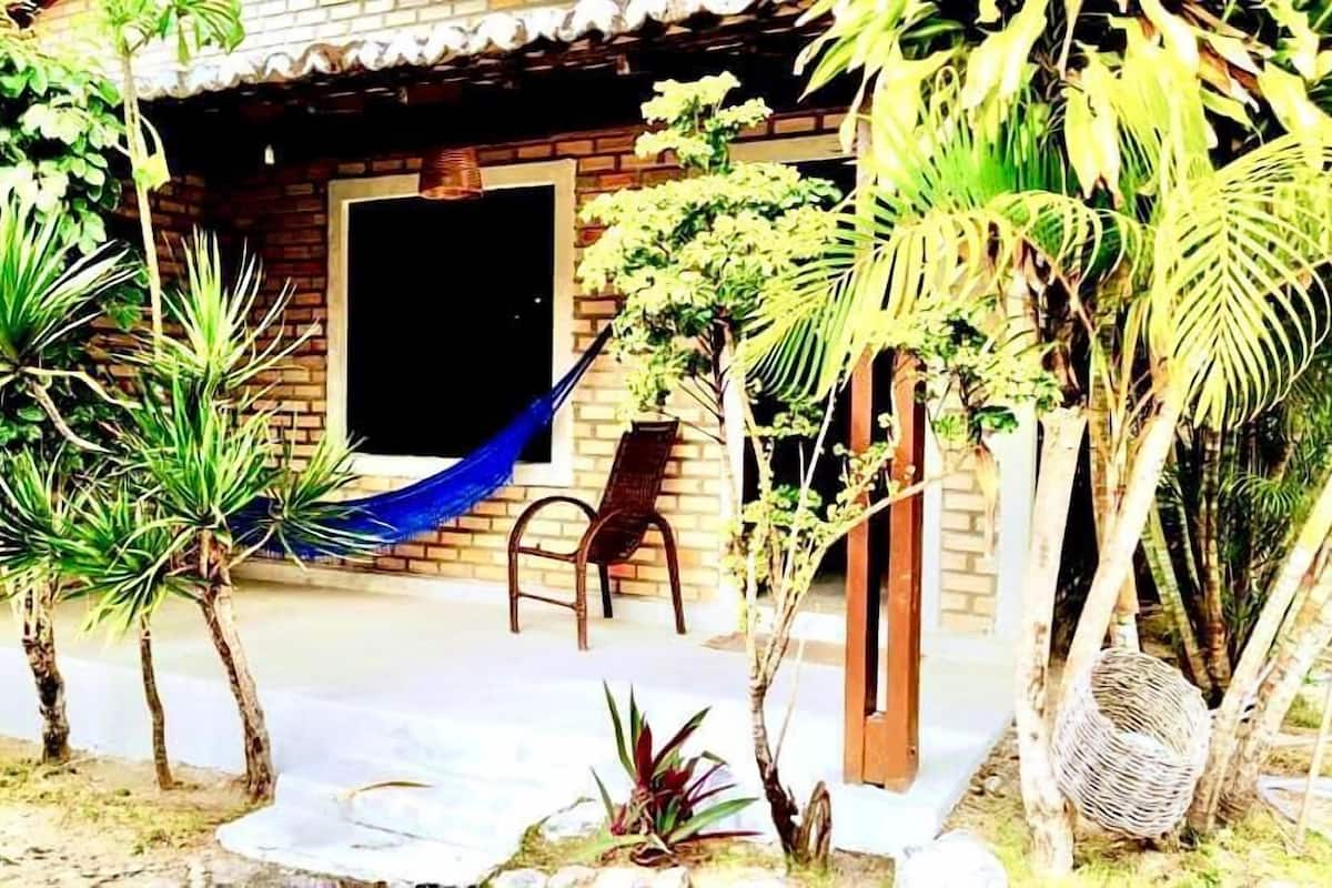 Il s’agit d’un bâtiment rustique à l’extérieur en briques, avec une porte en bois et un hamac. Plusieurs plantes en pot et un panier en osier se trouvent près de l’entrée.
