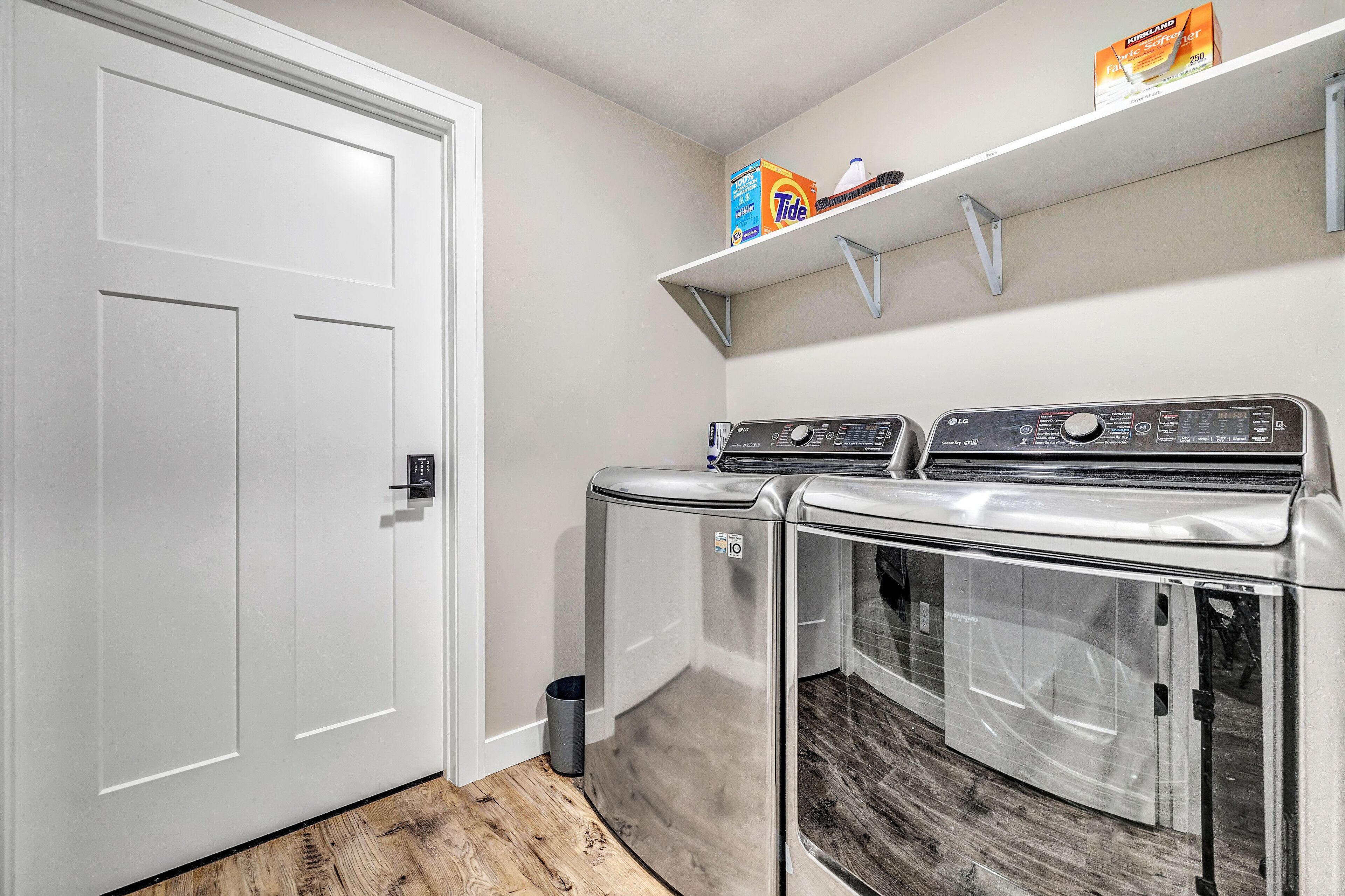 A laundry room with two front-loading washers and dryers, a white door, and a shelf above holding laundry supplies.