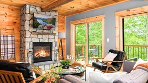 A cozy cabin interior with a stone fireplace, wooden ceiling, and large windows offering a view of the outdoors.