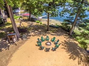 Un espace circulaire aménagé avec des chaises disposées autour d’un foyer central, situé sur une plage de sable, avec des arbres et une étendue d’eau en arrière-plan.