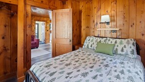 Quarto com painéis de madeira, uma cama, uma porta, uma luminária fixada na parede e vista para outro cômodo.