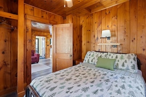 A wooden cabin bedroom with a bed, a nightstand, a wall-mounted lamp, and a door leading to another room.