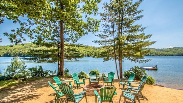 Un área de estar junto al lago con sillas y una chimenea, rodeada de árboles y vistas al lago.