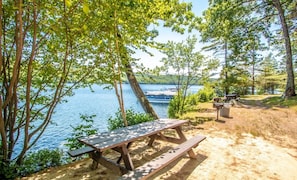 A picnic table and benches by a lake with trees and a boat dock in the background. - Squam Lake 3 bed 3 bath (Suite #7) (Holderness)