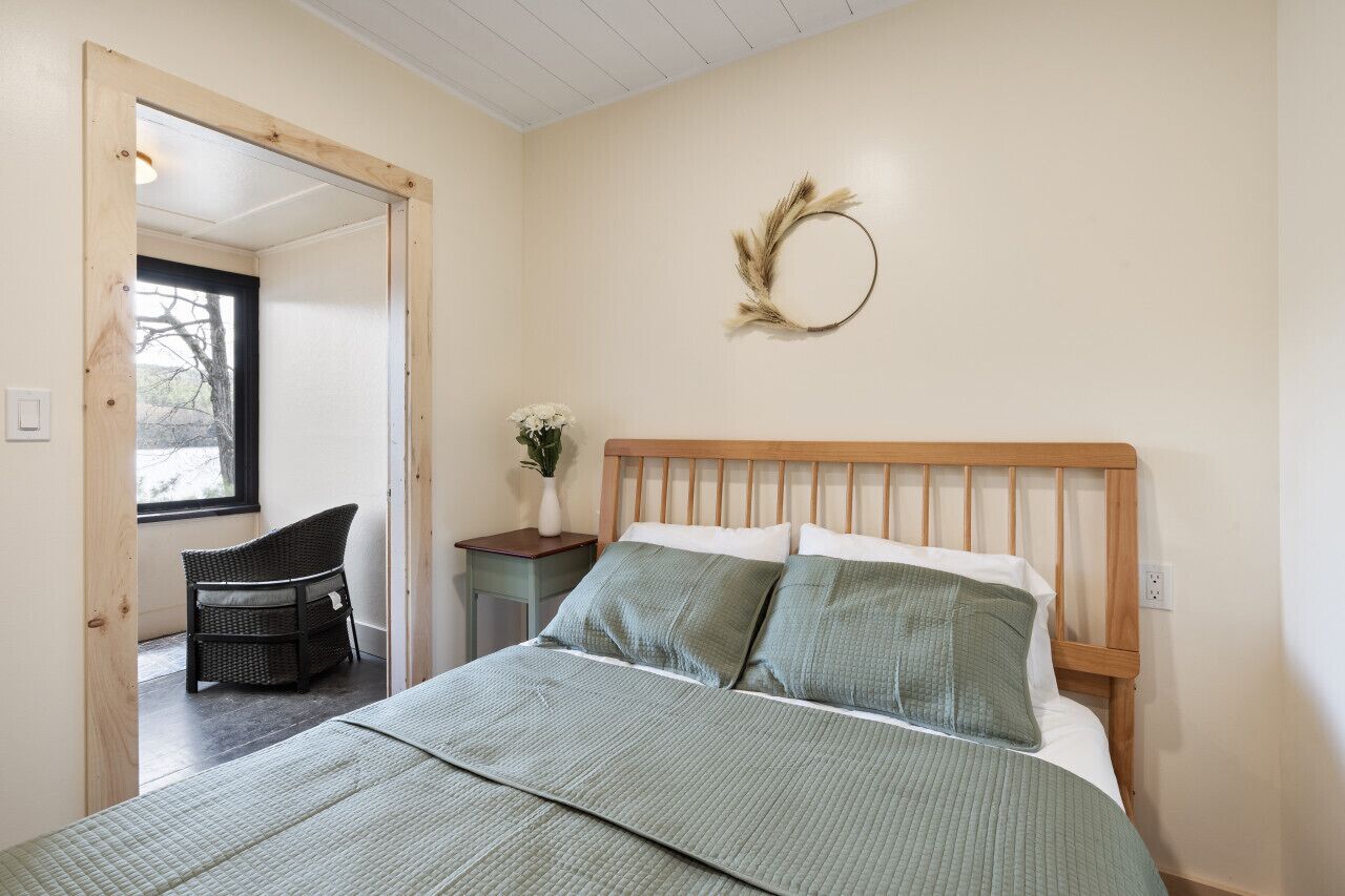 A bedroom with a wooden headboard, a green bedspread, a wicker chair, a small table with a vase, and a wall-mounted wreath.