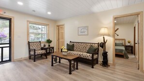 A cozy living room with a sofa, chairs, a coffee table, and a lamp. There is a door leading to another room and a window with a view of trees. - Waterfront 3 bed/3 bath on Squam (#1) (Holderness)