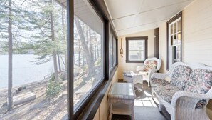 A cozy room with a large window overlooking a forest and a body of water. There is a wicker sofa, a small table, and a chair with floral cushions. - Moose Manor- Squam Waterfront (Holderness)