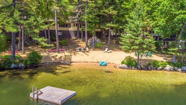 Un chalet au bord du lac, doté d’un quai, entouré d’une forêt dense et bordé d’une plage de sable où se trouvent des chaises longues.