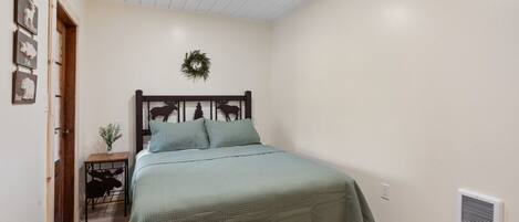 A bedroom with a bed, a small bedside table, a wall-mounted wreath, and a vent on the wall.