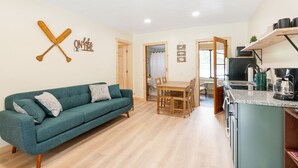 A living room with a teal sofa, wooden dining table, and kitchen area with a coffee machine. - Squam waterfront 2 bed 2bath (Suite 9) (Holderness)