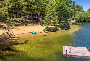 A lakeside property with a sandy beach, a wooden dock, and a row of lounge chairs.