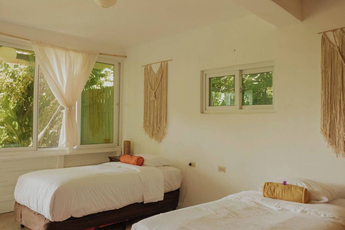 Een kamer met twee bedden, een raam met gordijnen en een wanddecoratie van macramé.