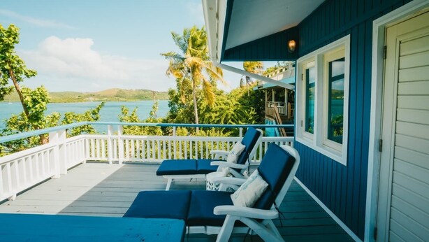 A covered deck with two lounge chairs and a small table, overlooking a body of water and palm trees. - AMAZING Ensenada Honda View @ MAREA 2-2 - Punta Aloe #3 (Culebra)