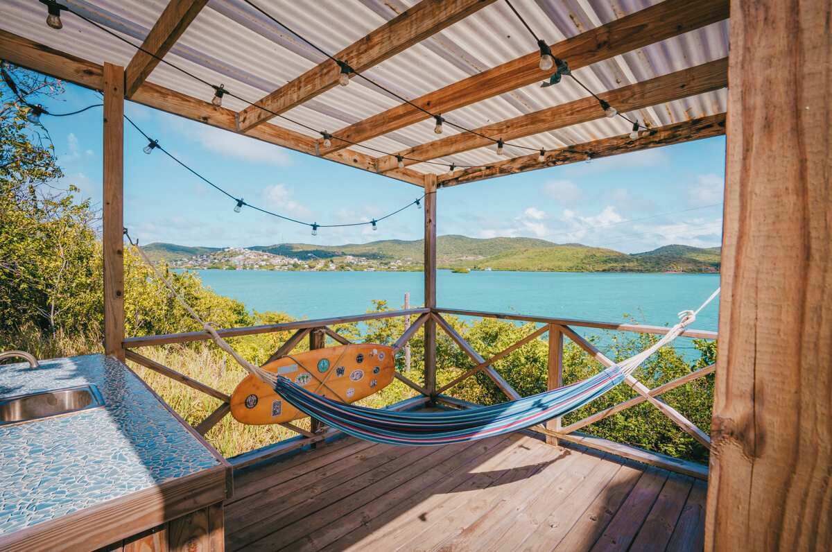 A wooden deck with a hammock and a table, overlooking a body of water and hills.