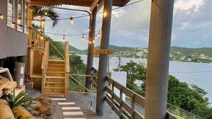 Un'area esterna coperta con una scala in legno che conduce a un livello superiore, luci decorative e vista su un corpo d'acqua e vegetazione.