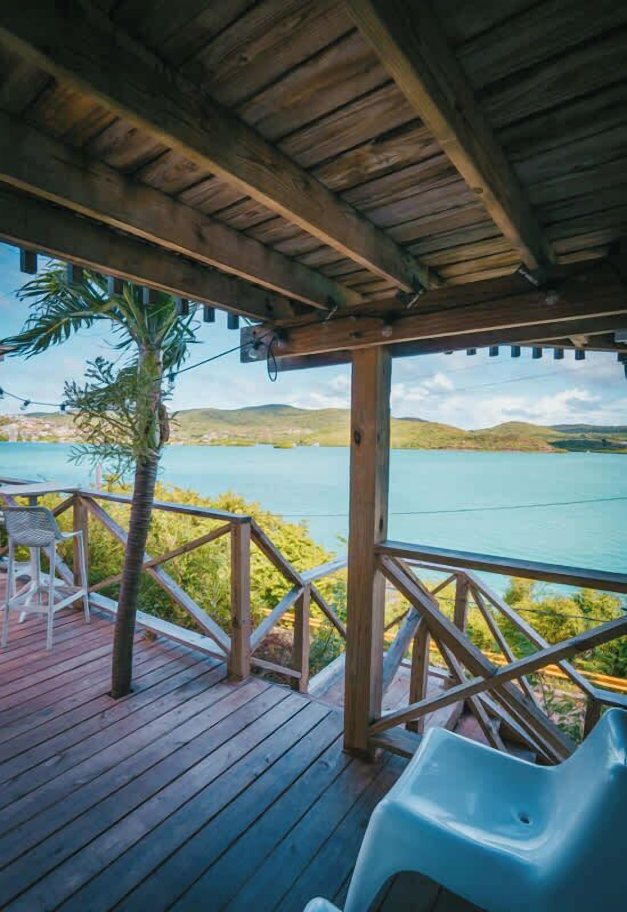 Une terrasse en bois dotée d’une balustrade, qui offre une vue sur une étendue d’eau et des collines.