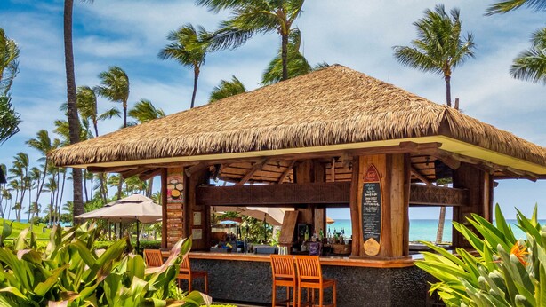 A beachside bar with a thatched roof, wooden chairs, and a menu board.