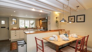 A kitchen with a wooden dining table set for four, white cabinets, a trash bin, and a window with blinds. - Kyte Cottage (Shipston on Stour)