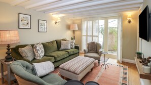 A cozy living room with a green sofa, a striped ottoman, and a small round table. There are two wall-mounted lamps and a framed picture on the wall. - Kyte Cottage (Shipston on Stour)