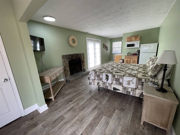 A bedroom with a fireplace, a bed with a patterned bedspread, a television, and a kitchen area with cabinets and a microwave.