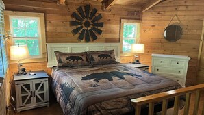 Un dormitorio tipo cabaña de madera con una cama que muestra un colcha con estampado de oso, un reloj de pared redondo y un mueble bajo con cajones.