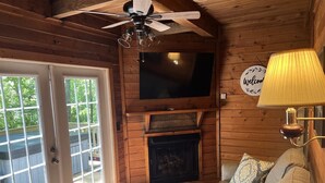 Interior acogedor de una cabaña con chimenea, un televisor montado encima, un sofá con cojines y un ventilador de techo.