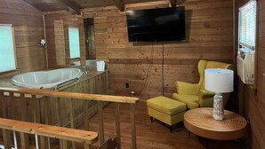 Interior de una cabaña de madera con una bañera de madera, un sillón amarillo, una mesa redonda con una lámpara y un televisor montado en la pared.