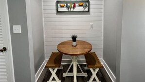 A corner room with a white wall, a wooden bench, a round table, and a framed artwork above the bench. - Rivers Edge 102 (Helen)