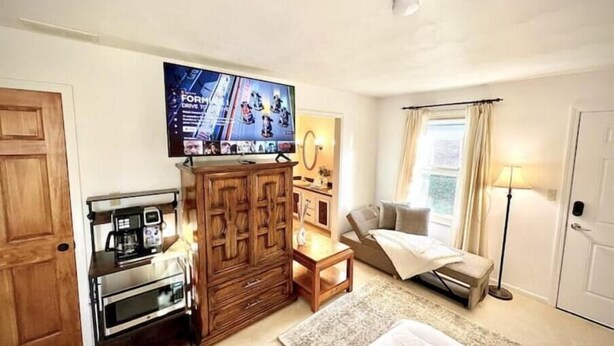 A hotel room with a wooden cabinet, a television mounted above it, a bed with a white comforter, a small bedside table, a floor lamp, and a window with curtains. - Neat & Tidy Private Entry Cozy Suite in Dayton Area with a Dedicated AC (Dayton)