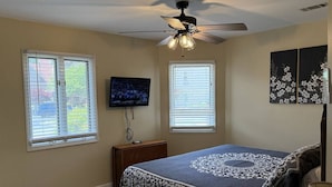 Un dormitorio con cama, ventilador de techo, dos ventanas con persianas, un televisor sobre un mueble bajo y un cuadro en la pared.